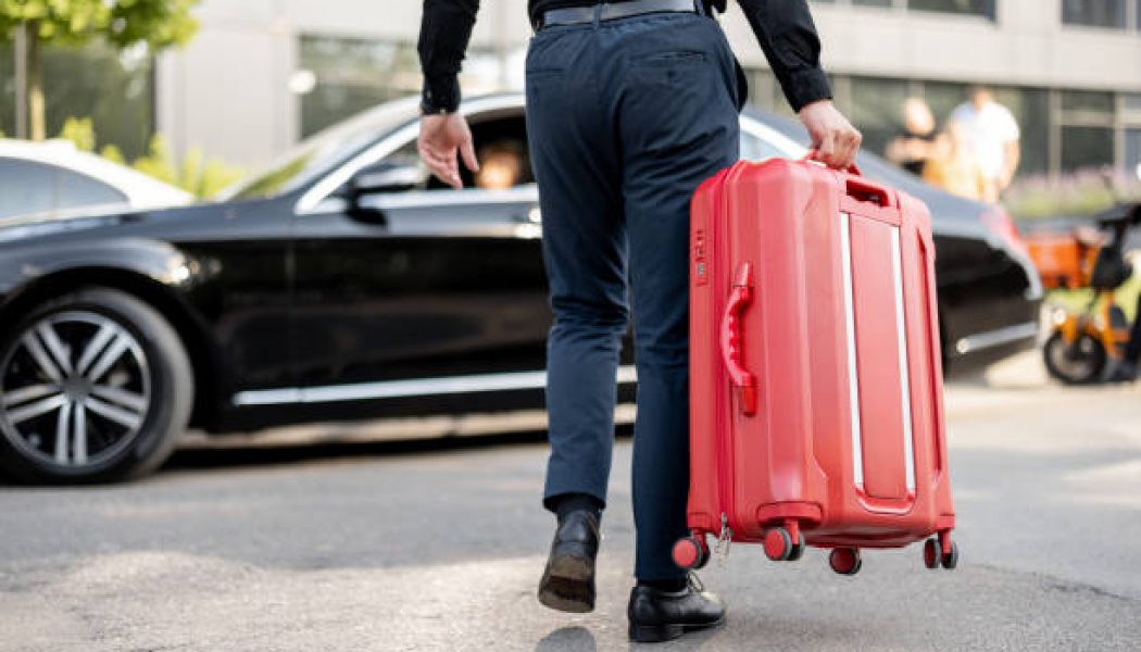 Man carries red suitcase to a car, cropped view from below. Concept of chauffeur service and business trips